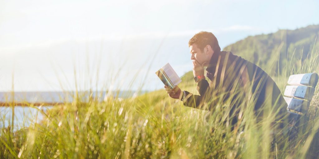 man reading a book thoughtfully, on a bench in grass by the shore.
