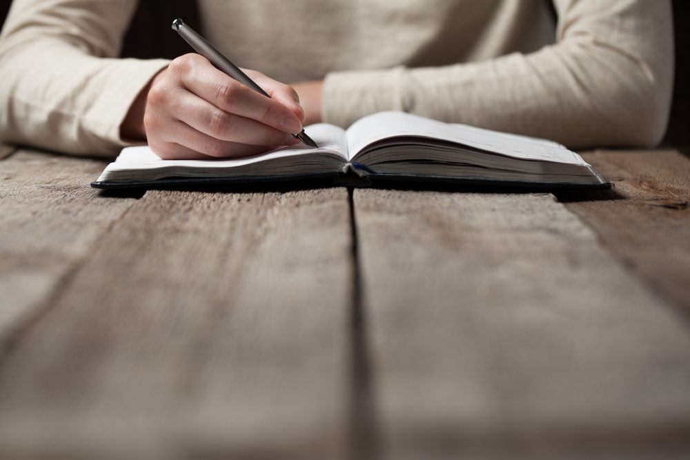 person writing with pen into journal on a wooden table