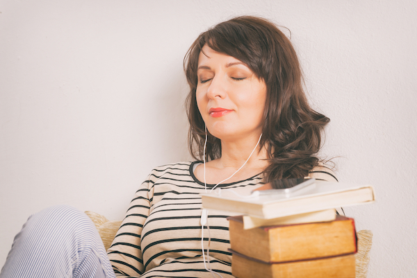 Woman listening an audiobook on smartphone sitting between piles of paper books.