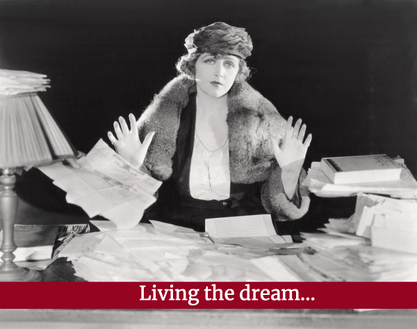 Elegant woman at a table swamped in papers, black and white, with words "Living the dream"