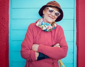 Olderwoman in bright sweater, sunglasses and hat, smiling.